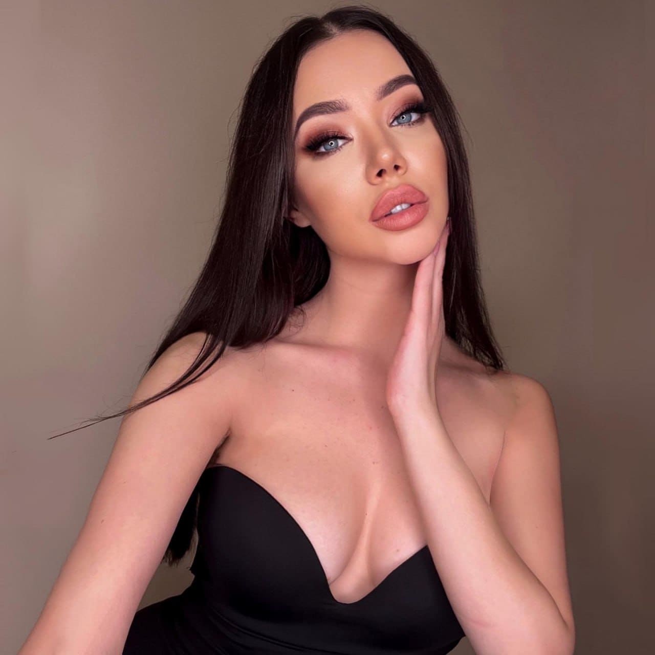 Милла в черном топе без бретелей с глубоким вырезом, рука у щеки, на фоне бежевой стены.