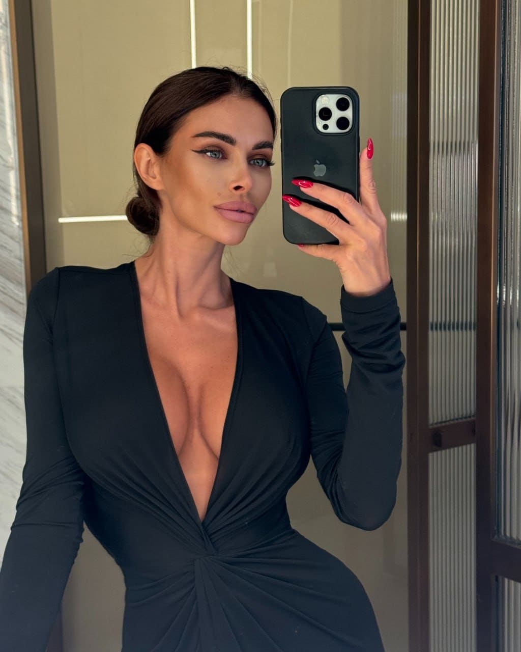 Нона в чёрном платье с глубоким вырезом делает селфи в зеркале на фоне стеклянной двери с вертикальными рёбрами.